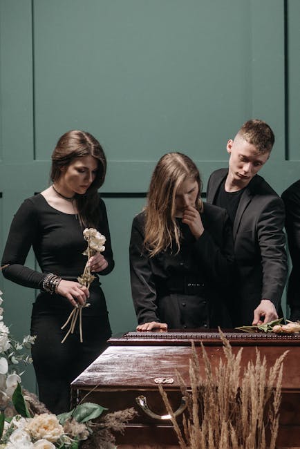 Three individuals standing beside a wooden casket, engaged in a somber moment during a funeral service. The woman on the left holds a small bouquet of white flowers, her expression reflective and subd