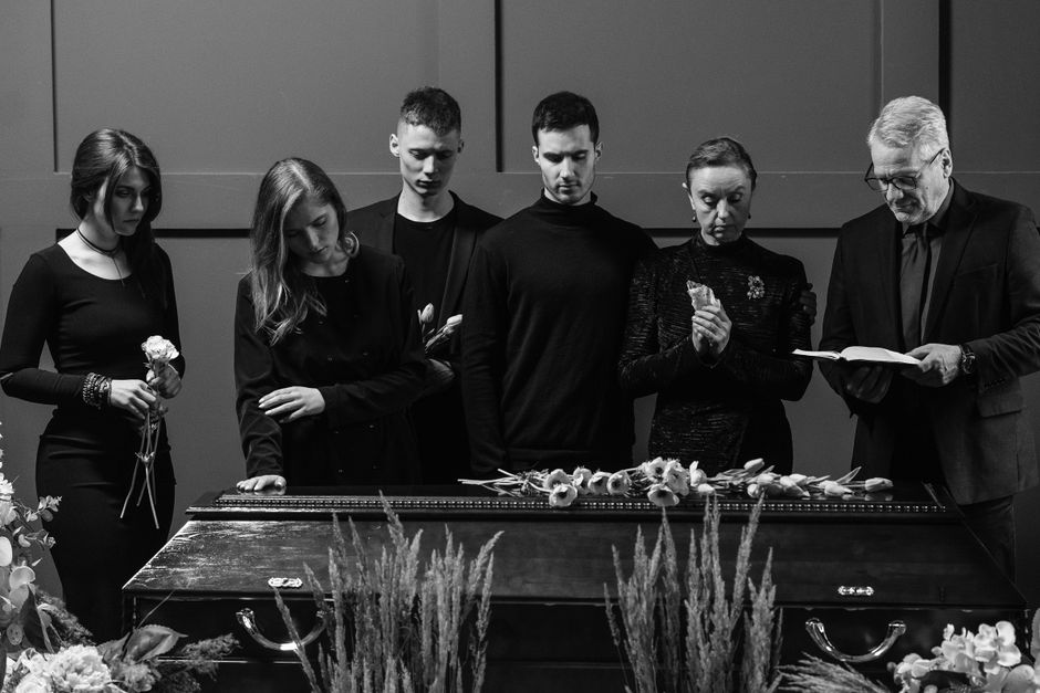 A black and white photograph depicting a group of six people gathered around a casket during a funeral service. The casket is adorned with a floral arrangement composed of delicate flowers, possibly r