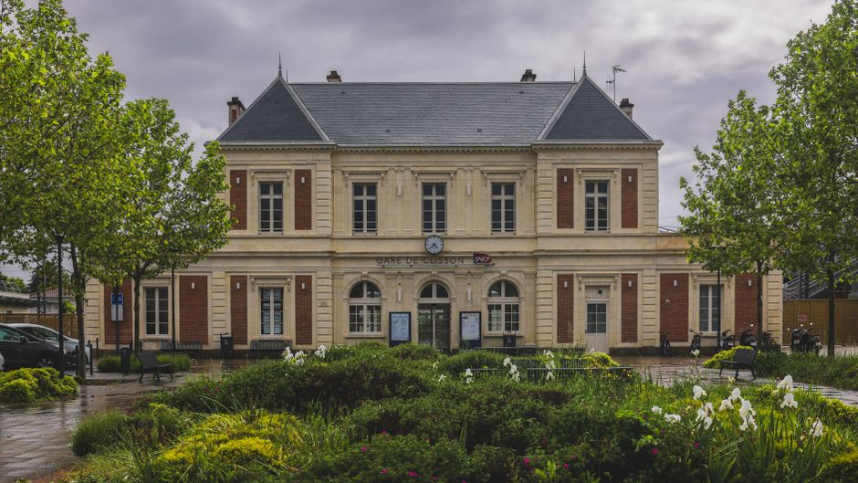 A historic train station building with a classic beige stone facade and tall arched windows, situated in a well-maintained garden area with vibrant green trees on either side. The station has a steepl