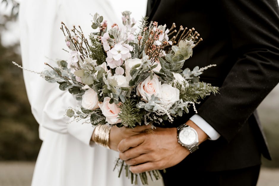 A couple holding a floral bouquet arrangement featuring soft pink roses, white peonies, and delicate sprigs of eucalyptus and other greenery, all arranged in a natural, loose style. The bride is weari