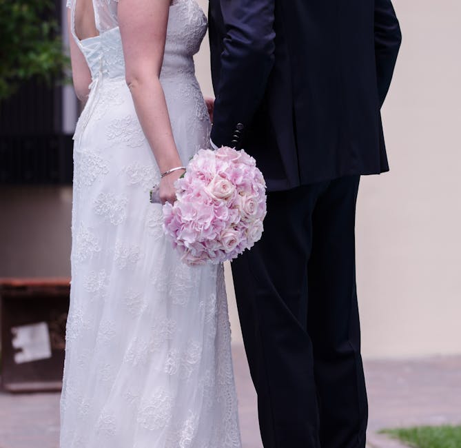 A bride and groom stand together, with the bride holding a round bouquet of pale pink roses and pink hydrangeas, featuring soft, fresh petals and green stems wrapped in a white ribbon. The bride wears