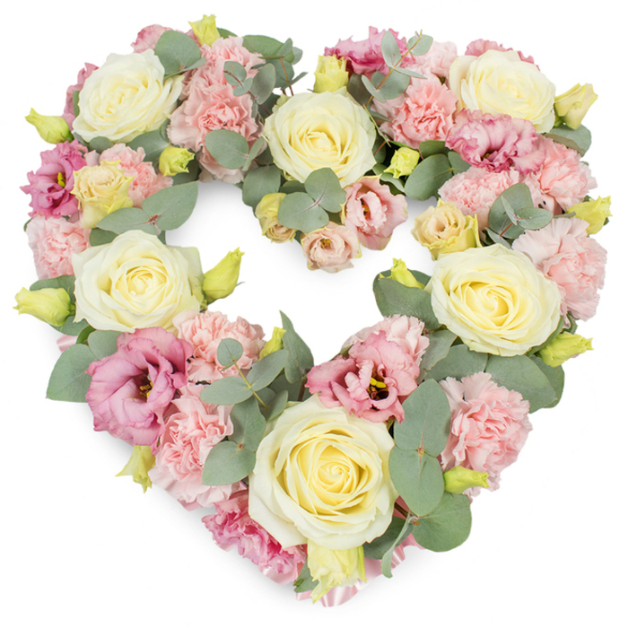 A tender, heart-shaped wreath designed as a funeral or memorial tribute is shown against a pure white background, its pastel palette and lush texture evoking a quiet calm reminiscent of an early morning walk through Hadley Common in Barnet. The heart is generously filled, every inch packed with blossoms so the form feels solid yet soft, like a comforting embrace. Dominating the design are creamy ivory to pale lemon roses, their velvety petals layered in precise spirals, catching subtle highlights that suggest freshness and careful handling. Between these focal roses, clusters of ruffled light pink carnations spill outward, each head composed of many frilled petals that add movement and a gentle, feathery depth. Deeper rose-pink and mauve lisianthus blooms are scattered thoughtfully through the arrangement, some more open, others still cupped, creating a play of shapes that draws the gaze around the heart. Tiny, unfurled yellow-green buds peek from the greenery, adding a sense of newness and quiet hope. Silvery eucalyptus leaves, with their smooth, rounded shapes and slightly matte finish, interlace among the flowers, cooling the colour scheme and defining the heart's outline with subtle curves. The overall mood is one of grace, remembrance, and steadfast affection, as though arranged by an experienced Barnet florist who often prepares tributes for services near Barnet Church or local crematoria. This heart wreath would be a fitting way to say a final, loving farewell, its soothing colours and textures offering comfort to family and friends gathered to honour a life well lived.